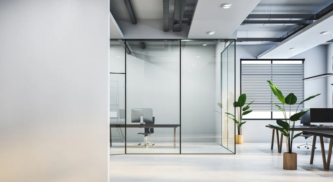 Modern commercial office space with glass partition walls, workstations, indoor plants, and open ceiling design