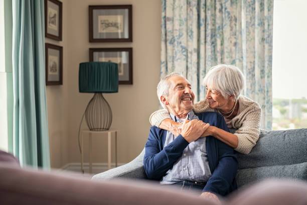 Happy senior woman embracing her husband at home while laughing together. Smiling wife hugging old man sitting on couch from behind. Joyful retired couple having fun at home while looking at each other with copy space. Love and unity concept.
