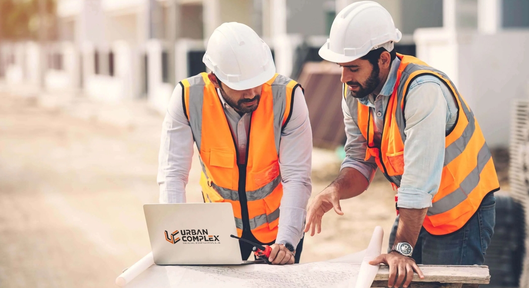Two construction engineers wearing white hard hats and orange safety vests reviewing blueprints